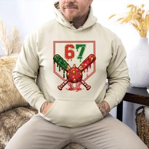 A graphic for apparel showing a large baseball ornament, complete with jingle bells, over two crossed baseball bats. The bats and the number 67 above them are styled like melting Christmas cookies with sprinkles. The design includes gingerbread men and snowflakes.