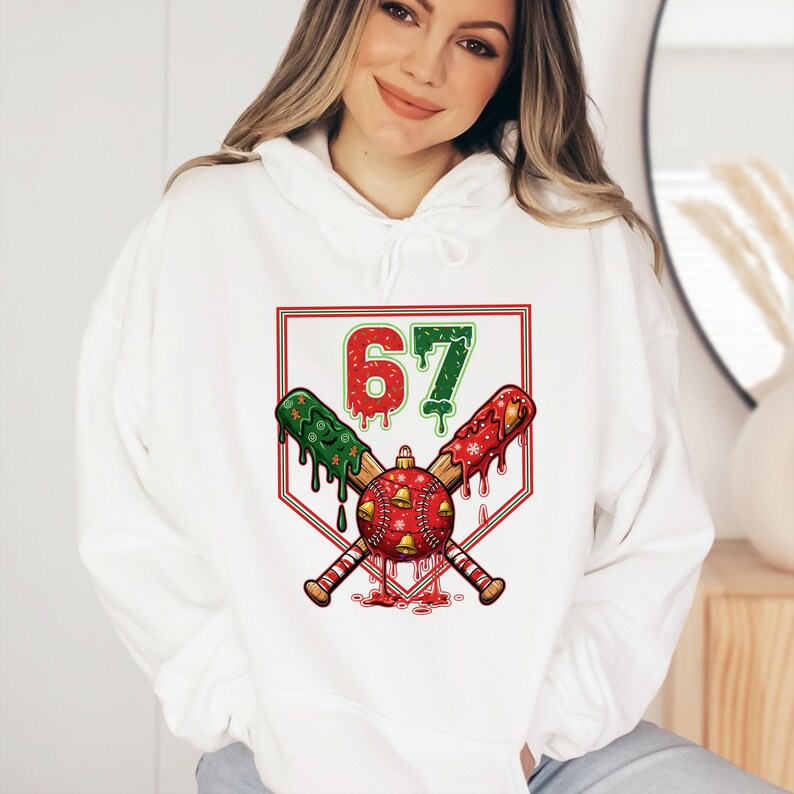 May include: White hoodie with a graphic design featuring the number 67 in red and green, crossed baseball bats, and a Christmas ornament. The bats are decorated with green and red patterns. The design is set within a red and white baseball diamond.