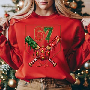 May include: Red sweatshirt with a baseball-themed graphic. The graphic features the number "67" in red and green, crossed baseball bats, and a baseball with bells. The design has a Christmas theme.