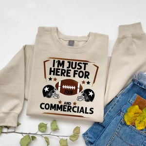 May include: A tan sweatshirt with a brown graphic that says "I'm just here for food and commercials" with a football and two football helmets.