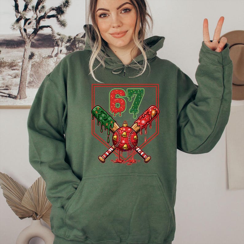 May include: Green hoodie with a graphic featuring crossed baseball bats, a baseball, and the number 67. The bats are decorated with red and green dripping designs. The baseball has red bells. The hoodie has a front pocket and a drawstring hood.