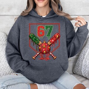 May include: Dark gray hoodie with a festive baseball-themed design. The graphic features crossed baseball bats, a baseball ornament with bells, and the number "67" in red and green. The design has a holiday theme.