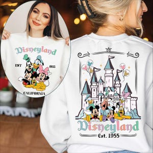 Puede incluir: Sudadera blanca con dise&ntilde;o de Disneyland. La parte delantera muestra la palabra "Disneyland" en colores pastel, con Mickey Mouse, el Pato Donald y otros personajes. La parte trasera muestra un castillo y personajes, con "Disneyland Est. 1955."