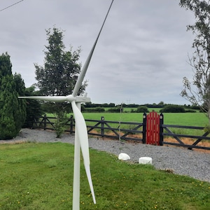 Op de afbeelding: Een witte windturbine met drie bladen is geïnstalleerd in een grasveld. De turbine bevindt zich in een woonwijk met een houten hek en een rode poort.