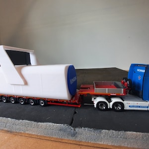 Puede incluir: Un objeto blanco cilíndrico con una tapa azul y un asa blanca se transporta en un remolque de plataforma plana tirado por un camión semirremolque azul y blanco. El objeto tiene la palabra "Vedder" impresa en la tapa azul. El camión semirremolque tiene la palabra "SCANIA" impresa en el lateral y la palabra "WHITTEN" impresa en la parte trasera.