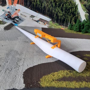 May include: A white model of a wind turbine blade is being lifted by a yellow crane. The blade is resting on a yellow metal frame with two supports. The model is on a gray and brown surface with green grass.