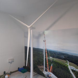 Könnte beinhalten: Ein weißes Modell einer Windkraftanlage mit drei Rotorblättern, vor dem Hintergrund eines im Bau befindlichen Windparks. Das Modell steht auf einem grauen Sockel, mit einer detaillierten Miniaturlandschaft im Vordergrund.