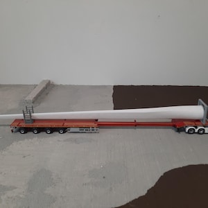 Puede incluir: Un camión semirremolque azul transportando una larga pala de turbina eólica blanca en un remolque de plataforma. La pala está asegurada al remolque con correas y el camión circula por una superficie de hormigón gris.