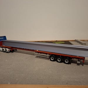 May include: A blue and white Scania truck towing a red trailer, transporting a large, grey steel beam. The model is on a grey surface, with a light-coloured background.