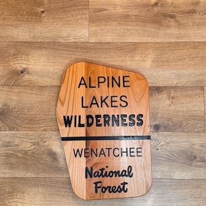 May include: Wooden sign with the text "ALPINE LAKES WILDERNESS WENATCHEE National Forest" in black lettering. The sign is a light brown color and has a unique shape. The background is a wooden surface.