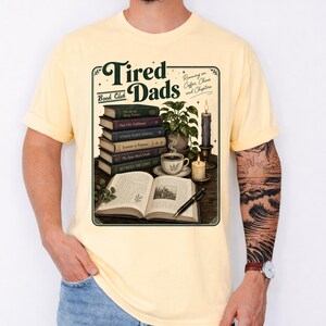 Peut inclure: T-shirt jaune pâle avec l'inscription "Tired Dads Book Club" et une illustration de livres, de café et d'une bougie. Le graphisme comprend les mots "Running on Coffee, Chaos, and Chapters."