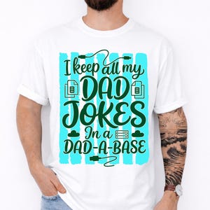 Op de afbeelding: Wit T-shirt met de tekst "I keep all my DAD JOKES In a DAD-A-BASE" in groen en turquoise. Het ontwerp bevat een gegevensserver, bestandspictogrammen en een wolkengrafiek.
