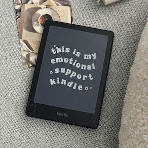 以下が含まれることがあります： 画面に「this is my emotional support kindle」と表示された黒いKindle電子書籍リーダーです。