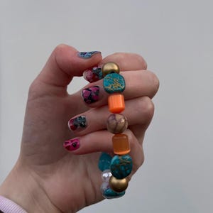 May include: A colourful beaded bracelet with turquoise, orange, gold, and white beads. The bracelet is being held by a hand with colourful nail polish.
