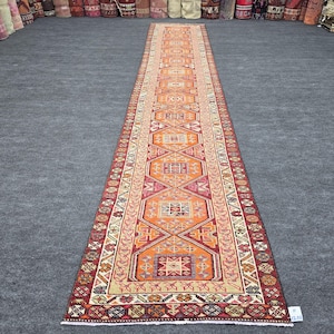Puede incluir: Una alfombra de pasillo larga y estrecha con un campo central de color naranja vibrante y motivos geométricos repetitivos de diamantes y cuadrados en tonos rojo, crema y azul oscuro. El borde muestra patrones intrincados en rojo intenso.