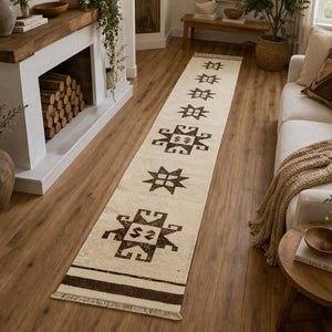 May include: A long, rectangular rug with a cream-colored background and dark brown geometric patterns. The rug features a repeating star and stylized square design. The rug is placed on a hardwood floor in a living room setting.