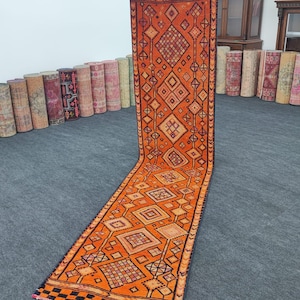 May include: An orange runner rug with a geometric pattern. The rug features diamond and square shapes in shades of orange, black, and beige. The rug is displayed on a gray surface, showcasing its length and design.