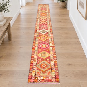 Puede incluir: Una alfombra de pasillo larga y estrecha con un patrón geométrico vibrante y repetitivo en tonos rojo, naranja, amarillo y crema. El diseño consiste en una serie de medallones en forma de diamante bordeados por intrincados motivos florales y lineales.