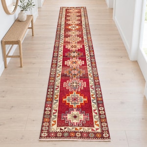 Puede incluir: Una alfombra de pasillo larga y estrecha con una base de color rojo vibrante y un patrón geométrico repetitivo en tonos crema, naranja, azul y gris. La alfombra tiene un borde decorativo con pequeños motivos florales y flecos en el extremo.
