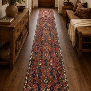 May include: A long, rectangular runner rug featuring a deep red background with intricate geometric patterns in shades of blue, cream, and dark brown. The rug has a symmetrical design with repeating motifs and a fringed edge at the bottom end.