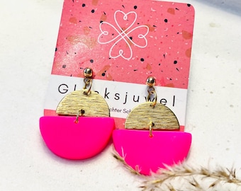 Puede incluir: Un par de pendientes de color rosa y dorado. Los pendientes están hechos de dos piezas: un semicírculo de acrílico rosa y un semicírculo de metal dorado. El metal dorado tiene un acabado texturizado. Los pendientes están en una tarjeta rosa con el texto "Glücksjuwel" y "chchter Sch".