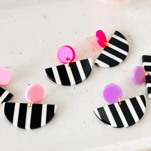 Könnte beinhalten: Geometrische Ohrringe mit einem halbkreisförmigen Design mit schwarz-weißen Streifen, kombiniert mit einem kleinen, runden Akzent in Rosa oder Lila. Die Ohrringe haben goldfarbene Stifte und sind vor einem weißen Hintergrund platziert.