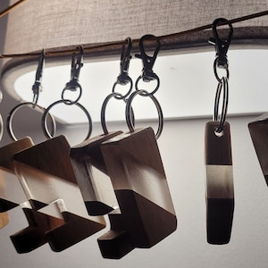 May include: A collection of wooden keychains with geometric designs. Each keychain features a combination of light wood, clear resin, and dark brown accents. The keychains are attached to silver-colored metal clasps and key rings.