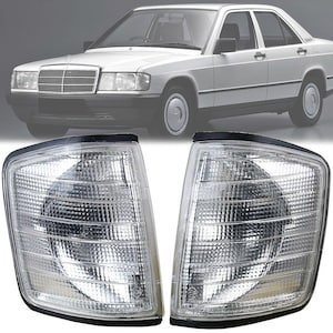 Puede incluir: Un par de luces de señalización delanteras blancas transparentes para un Mercedes-Benz 190E. Las luces tienen una carcasa de plástico negro y una lente transparente con un patrón de rejilla.