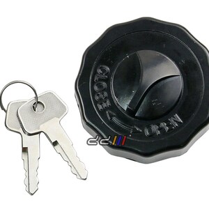 Op de afbeelding: Een zwarte tankdop met een sleutelgat en de woorden "OPEN" en "CLOSE" erop gedrukt. Twee zilveren sleutels zijn inbegrepen.