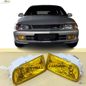 May include: A pair of yellow fog lights for a car. The lights are rectangular and have a white plastic housing. The lights are mounted on a white surface.