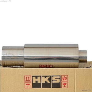 Pode incluir: Um silenciador de escape HKS prateado em uma caixa de papelão com o logotipo HKS impresso no lado.