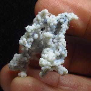 May include: A close-up of a mineral specimen featuring a cluster of small, round, white and light blue formations. The mineral has a textured, porous appearance, with some areas appearing translucent. The background is dark.