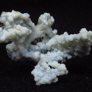 May include: A close-up of a mineral specimen with a complex, branching structure. The mineral has a pale blue and white color, with a textured, almost bubbly appearance. Fine, hair-like crystals are visible throughout the structure, set against a black background.