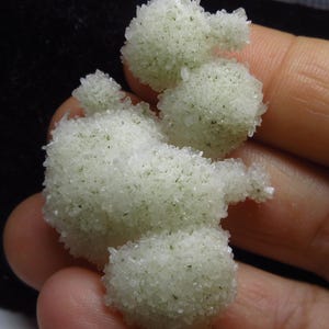 Könnte beinhalten: Eine Ansammlung von blassgrünen, kugelförmigen Mineralformationen mit einer kristallinen, texturierten Oberfläche. Das Exemplar weist mehrere abgerundete, blasige Auswüchse auf, die durchscheinend und funkelnd wirken, mit feinen dunklen Einschlüssen.