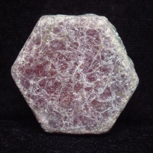 May include: A hexagonal, rough-cut ruby gemstone. The stone is a deep reddish-purple color with a network of white veins throughout. The gemstone is set against a black background.