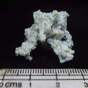 May include: A light blue mineral specimen with a cluster of small, rounded formations. The mineral has a textured, bubbly appearance and is set against a black background. A ruler with centimeter markings is at the bottom.