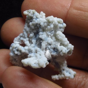 May include: A close-up of a mineral specimen held in a hand. The mineral has a light blue and white color with a bubbly texture. Fine, white, hair-like structures are visible throughout the specimen.