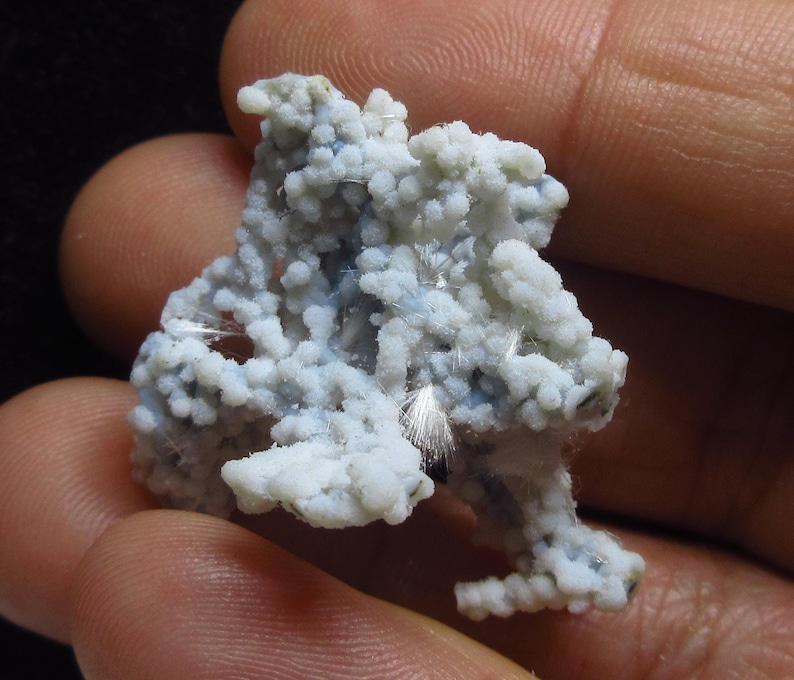 May include: A close-up of a mineral specimen held in a hand. The mineral has a light blue and white color with a bubbly texture. Fine, white, hair-like structures are visible throughout the specimen.