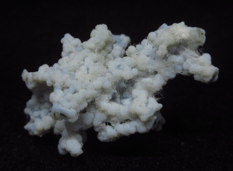 May include: A close-up of a mineral specimen with a light blue and white color. The mineral has a clustered, textured appearance, with small, rounded formations. Fine, white, hair-like structures are visible throughout the specimen.