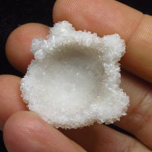 Könnte beinhalten: Ein kleines, weißes, kristallines Mineralexemplar mit einer hohlen, schalenartigen Mitte, die mit winzigen, funkelnden, durchscheinenden, quarzähnlichen Spitzen bedeckt ist. Der Cluster hat eine unregelmäßige, runde Form.