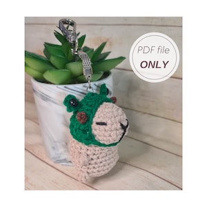 May include: Crocheted amigurumi keychain of a capybara wearing a green frog hat. The keychain is attached to a silver metal ring.