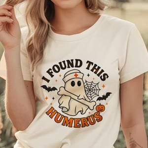 May include: Cream-colored t-shirt with a Halloween-themed graphic. The design features a ghost wearing a nurse's cap and holding a bone, with the text "I FOUND THIS HUMERUS". Includes bats, a spiderweb, and a pumpkin.