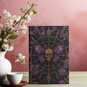Hardcover journal featuring an ornate gothic design of a golden skull with purple butterfly wings at the center of a golden spiderweb. Surrounded by rich purple flowers, butterflies, and intricate details, all set against a dark, swirling background.