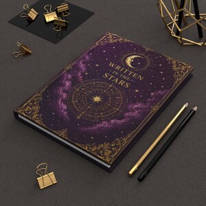 May include: A dark purple hardcover journal with gold foil celestial illustrations, including a crescent moon, stars, and a compass design. The cover features the text "WRITTEN IN THE STARS" in gold lettering, surrounded by ornate gold filigree borders.