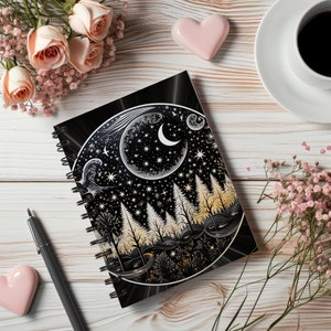 May include: A black spiral notebook with a moon and forest design. The moon is white and crescent shaped. The forest is black and white with a gold outline. The notebook is on a white wooden surface.