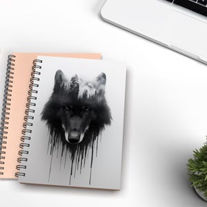 May include: A black and white notebook with a wolf's head design. The wolf's head is made up of trees and a forest scene. The notebook has a spiral binding and a pink cover.