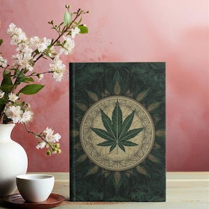 Op de afbeelding: Een groen notitieboek met een cannabisbladontwerp, 14,6 cm x 20,3 cm, en 150 gelinieerde pagina's. Het notitieboek heeft een matte afwerking. Een witte vaas met bloemen en een kleine witte kopje staan ook in de afbeelding.