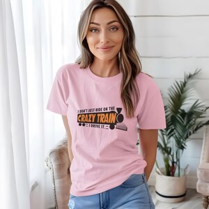 Puede incluir: Camiseta rosa con un gráfico negro y naranja que dice "No solo me monto en el tren loco, lo conduzco."