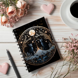 May include: A black spiral bound notebook with a gold and black illustration of a moon, stars, and two black cats. The illustration is surrounded by a gold circle with a gold border.