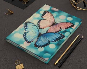 May include: A hardcover journal featuring a butterfly design. The cover displays three butterflies with blue and pink wings against a teal background with bokeh lights. Gold and black pencils and gold binder clips are also present.